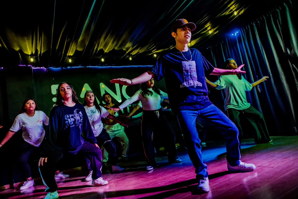 Instructor de hip hop guiando la coreografía con el grupo detrás, en el salón iluminado de colores