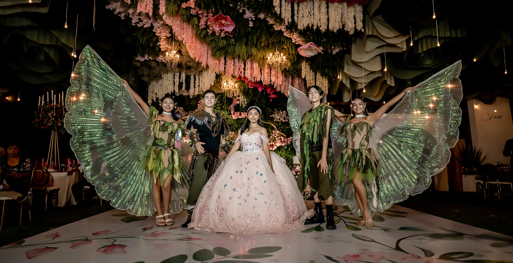 Quinceañera con vestido rosa pálido rodeada por cuatro bailarines de Dance Central con alas iridiscentes, bajo un techo de flores