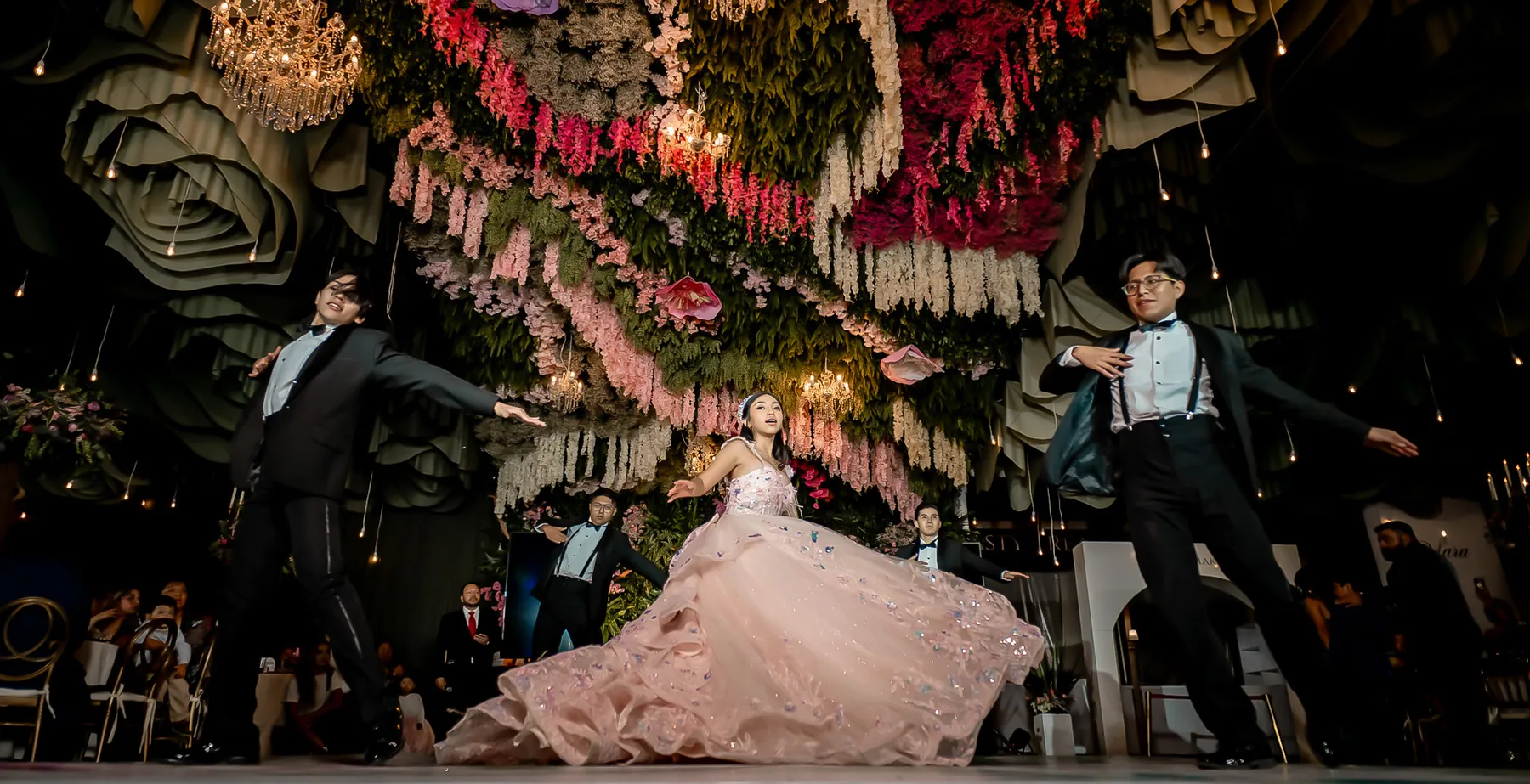 Quinceañera bailando el vals acompañada de dos chambelanes de traje en una pista decorada con flores
